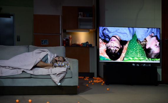 En man ligger under en filt i en soffa bredvid en tv som visar två personer. Orange bollar ligger utspridda på golvet.