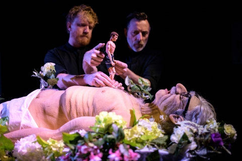 En man ligger ner omgiven av blommor, på hans bröst går en actionfigur som två andra män håller i. 