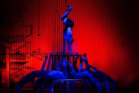 Framför den rött upplysta bakgrunden syns 4 akrobater i blått ljus. En av dem står på hände ovanpå ett bord som de övriga akrobaterna lyfter upp. Akrobaterna som fungerar som bordsben ligger på rygg ovanpå en plattform och sträcker sina armar rakt upp i vädret för att stötta denna balansakt 