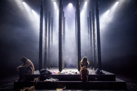 En dramatisk bild från föreställningen Closer där två skådespelare sitter vända från varandra på en upphöjd svart scenplattform. Scenen är upplyst av flera strålkastare ovanifrån som skapar dramatiska ljusstrålar genom teaterdimma. Mellan personerna ligger några föremål utspridda, inklusive väskor, flaskor och vad som ser ut som klädesplagg. Personerna bär bruna kläder och sitter i nedböjda, sorgsna positioner. 