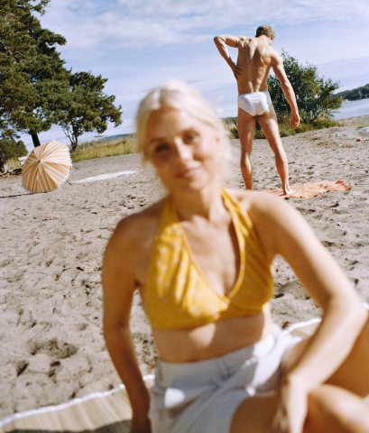 Foto: Felix Swensson Kvinna i gult bikinilinne sitter suddigt i förgrunden på en sandstrand, medan en man i vita badkläder står i bakgrunden vid vattnet.