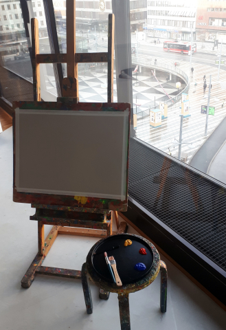 Staffli med en tom duk och en målarpall med tre färgdroppar och penslar, vid ett fönster med utsikt över en stadsmiljö.