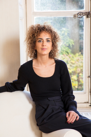 Foto: Francesca Mantovani © Gallimard Yngre kvinna med lockigt hår och svart topp sitter inomhus framför ett fönster.