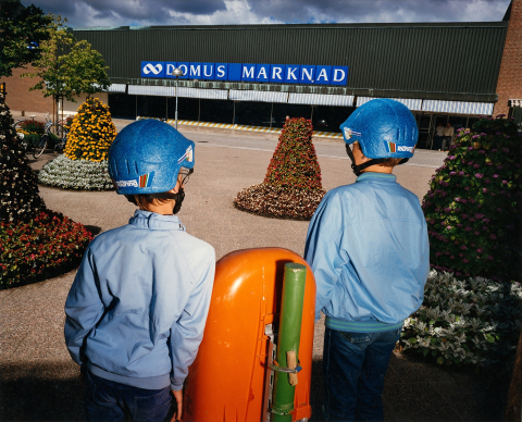 © Lars Tunbjörk, Maud Nycander, Stiftelsen Lars Tunbjörk Två pojkar i blå hjälmar och jackor står vända mot varuhuset Domus Marknad, omgivna av blomstertorn.