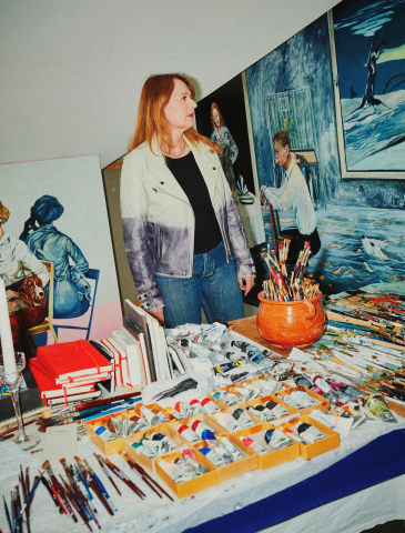 Konstnären Meta Isæus-Berlin står vid bord med penslar, färgtuber och målningar i en ateljé.