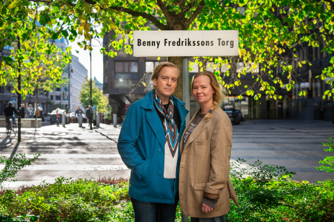 Foto: Leonard Stenberg Två vuxna personer står framför skylten "Benny Fredrikssons Torg" i en stadsmiljö med träd och byggnader.