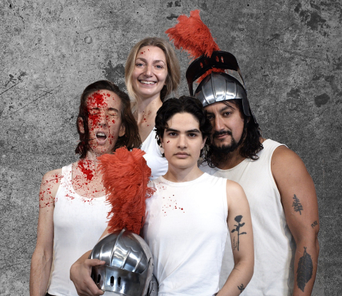 Fyra vuxna i vita linnen, tre med blodstänk, två bär romerska hjälmar med röd plym; betongvägg i bakgrunden.