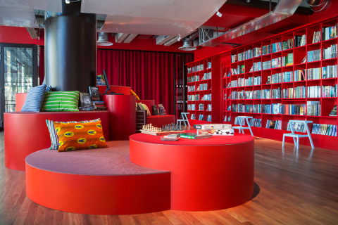 Modernt bibliotek med röda bokhyllor, rund sittplats med kuddar, sällskapsspel och schackbräde på bordet.