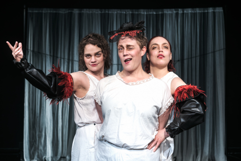 Tre kvinnor i vita underkläder, två med svarta ärmar med röda fjädrar, poserar framför ett draperi på en scen.