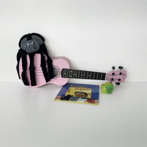 Rosa ukulele med en svart spindelgosedjur ovanpå, barnbok och grön snigelleksak framför.