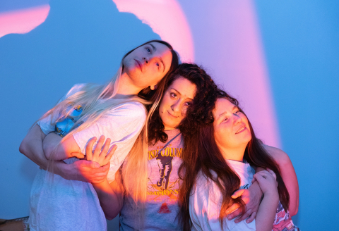 Tre unga kvinnor med långt hår står tätt ihop framför en blå vägg med rosa och lila ljus.