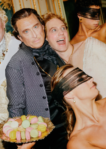 Fem personer i barockinspirerade kläder, två med ögonbindlar, en håller ett fat med färgglada macrons.