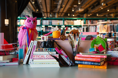 Bord fullt av färgglada konstmaterial, böcker, penslar, kamera och en rosa uggla i ett bibliotek.