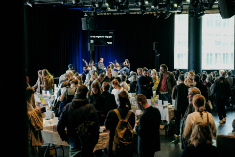 Många vuxna besökare bläddrar bland böcker på bord; en man och en kvinna sitter på scen i bakgrunden.