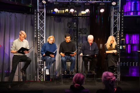 Sex personer, tre män och tre kvinnor i olika åldrar, sitter på pallar på scen och läser ur svarta manus.