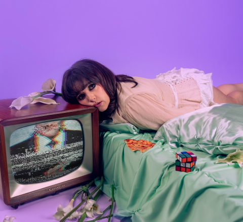Kvinna ligger på säng med huvudet mot en tv med brus; pizza, Rubiks kub och blommor runtom i scen med lila bakgrund.