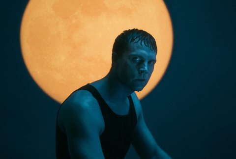 Person i svart linne sitter i blått ljus framför en stor orange cirkel som liknar en sol eller måne. Bilden har dramatisk kontrast och en mörk, filmisk stämning.