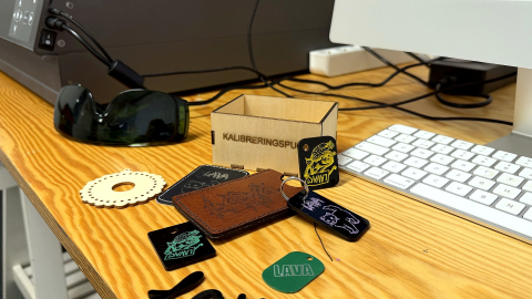 Laserskurna och graverade föremål i trä, läder och plast ligger på ett arbetsbord bredvid ett tangentbord.