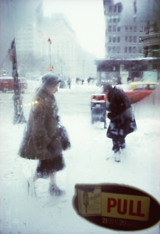 Två personer går genom ett snöigt stadslandskap i kraftigt snöfall, fotograferade genom en glasdörr med skylten ”PULL”.