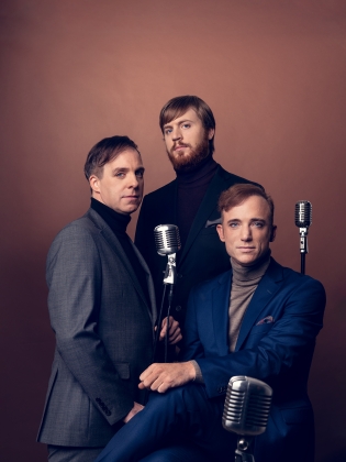 Albin Flinkas, Fredrik Meyer och Fabian Fredriksson i kostymer mot en brun bakgrund med äldre mikrofoner