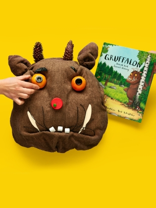 Ett monsterliknande huvud med orange ögon, röd näsa och betar, i bakgrunden boken Gruffalon. 