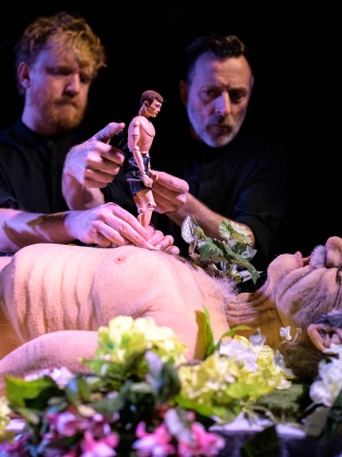 En man ligger ner omgiven av blommor, på hans bröst går en actionfigur som två andra män håller i. 