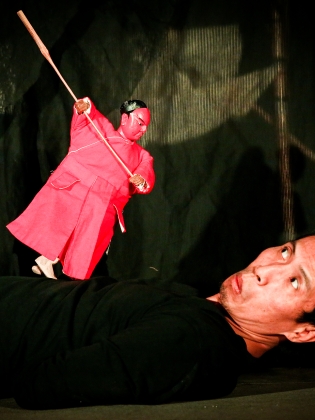 En man som ligger ner håller i en docka som riktar en lång stav mot honom. 