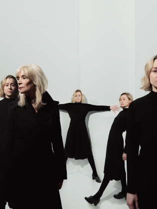En kvinna i blont hår och svarta kläder håller ut armarna åt sidan, runt henne står fler kvinnor klädda i svart. 