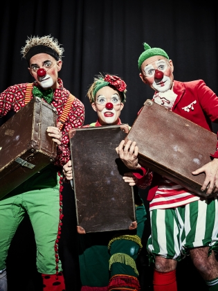 Svart bakgrund. Tre personer med iklädda clownkostym och röd clownnäsa. Alla tre personer håller i varsin koffert.