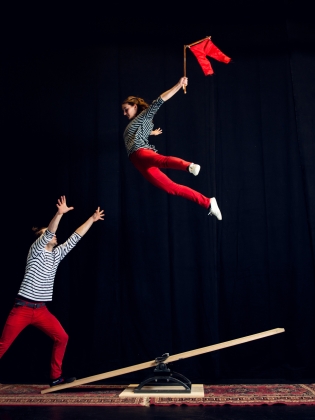 Två cirkusartister på en språngbräda. Den ena flyger i luften med en flagga i handen
