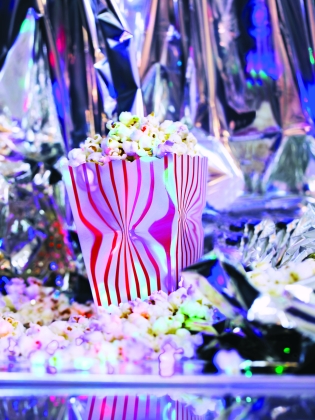 En pappersbägare i holografiska neonfärger fylld med popcorn står på en bädd av popcorn. Bakgrunden är holografisk och silverfärgad