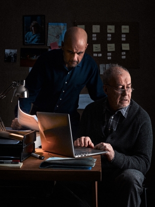 Två män vid ett skrivbord, de går igenom olika papper och arbetar med en dator. 