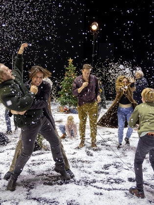 Det snöar på en teaterscen där flera skådespelare leker i snön. 