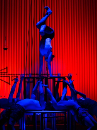Framför den rött upplysta bakgrunden syns 4 akrobater i blått ljus. En av dem står på hände ovanpå ett bord som de övriga akrobaterna lyfter upp. Akrobaterna som fungerar som bordsben ligger på rygg ovanpå en plattform och sträcker sina armar rakt upp i vädret för att stötta denna balansakt 