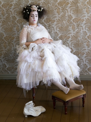 Gaby Muñoz sitter på en stol iklädd en bröllopsklänning. Fötterna är uppslängda på en pall och skorna ligger intill på golvet. Gaby är vitmålad i ansiktet och har röda läppar och höjda ögonbryn. Gaby har vita blommor i håret . Rummet är tomt, bakom henne- en klassiskt mönstrad tapet 