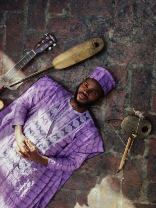 Ebo Krdum iförd en lila dräkt liggandes på ett stengolv med musikinstrument runt sig