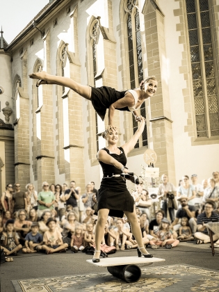 Två cirkusartister som utför akrobatik framför en kyrkbyggnad med publik
