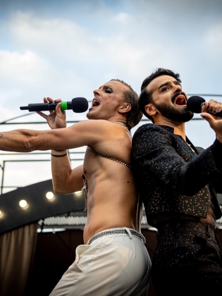 Två musikalartister står rygg mot rygg och sjunger med armarna utsträckta