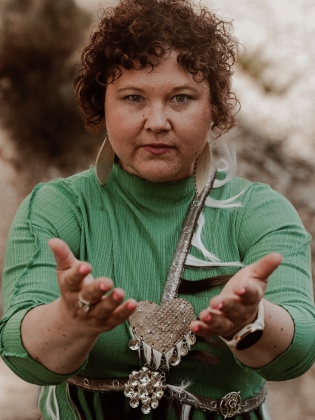 Sara Ajnnak i en grön klädsel med samisk utsmyckningar. Hon håller händerna utsträckta framför kroppen