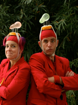 Två rödklädda cirkusartister med vattenballonger på huvudet