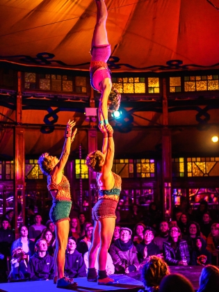Tre cirkusartister på scen. En av akrobaterna står på händer uppe på en annan akrobats händer. 