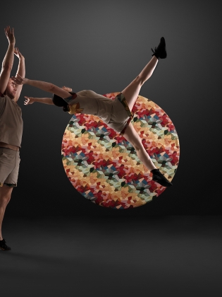 Två akrobater på en upplyst scen. Den ena kastas ur den andras armar och bakom dem är ett runt objekt med färgglatt mönster.