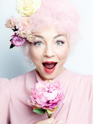 Sara Jangfeldt med rosa kläder, peruk och blommor i håret. Hon är glad och håller en stor blomma i handen