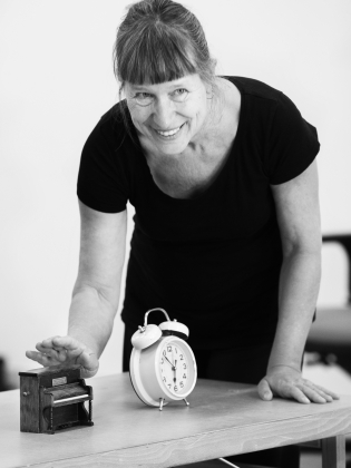 En kvinna står vid ett bord med en väckarklocka och ett miniatyrpiano på. 