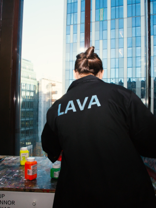 Person i svart jacka med texten "LAVA" målar vid ett bord med fönsterutsikt över stad.