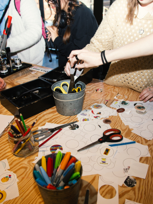 Grupp vid ett bord skapar klistermärken, med saxar, pennor och papper utspridda.
