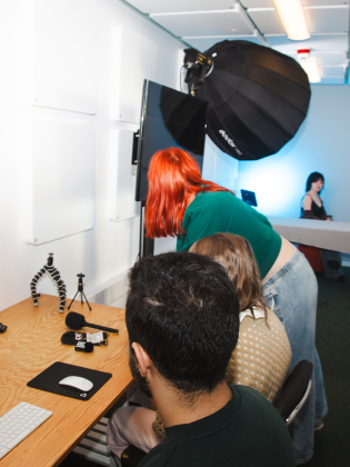 Flera personer i en studio med dator, fotoutrustning och lampor; en person fotograferar i bakgrunden.