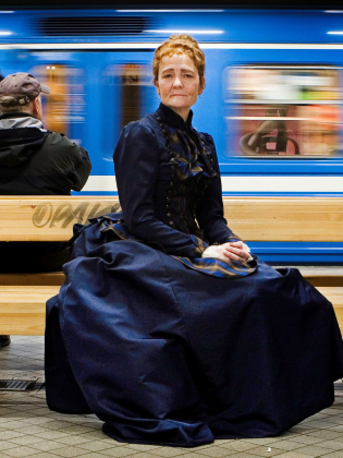 Ann Petrén i en gammeldags klänning sittandes på en bänk på en tunnelbaneperrong