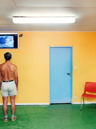 Skjortlös man i shorts står i färgglatt rum och tittar på en liten TV ovanför en blå dörr.