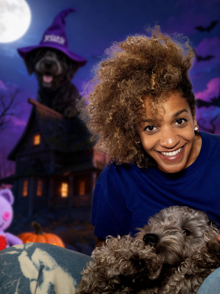 Kvinna med lockigt hår i blå tröja sitter med en grå hund i knät, framför en halloweenbakgrund med måne och pynt.